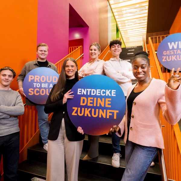 Auszubildende stehen auf einer Treppe und schauen in die Kamera mit Schildern mit Aufdruck "Wir gestalten Zukunft" und "Entdecke deine Zukunft"