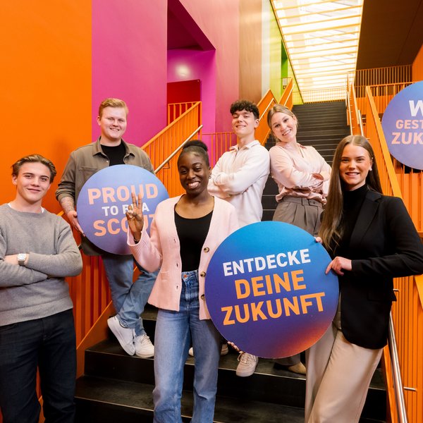 Auszubildende stehen auf einer Treppe und halten Schilder mit der Aufschrift "Entdecke deine Zukunft" und "Wir gestalten Zukunft".